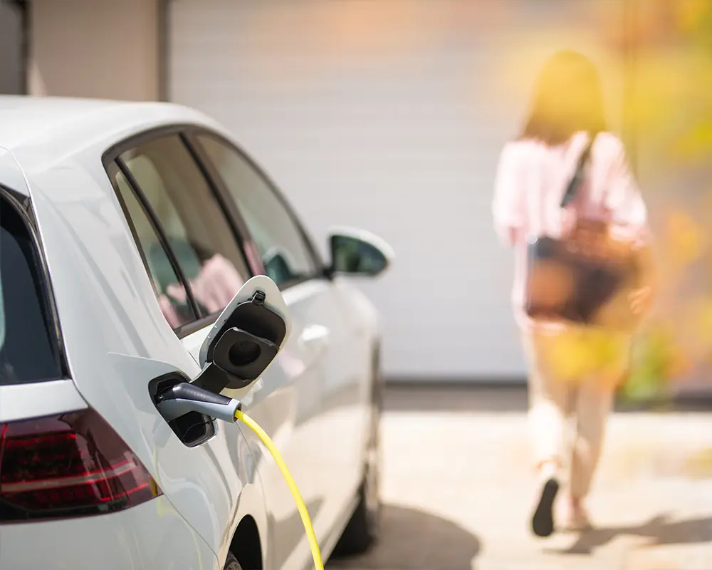 White electric car charging in driveway while woman walks away.